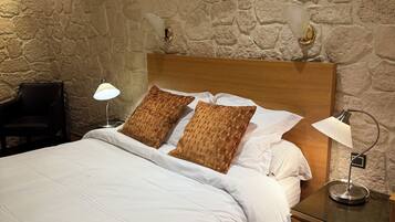 Un lit bien fait, avec du linge de lit blanc et deux coussins à motifs, une lampe de chevet et une table de chevet dotée d’un tiroir. La pièce est agrémentée d’un mur en pierre et d’une tête de lit en bois.