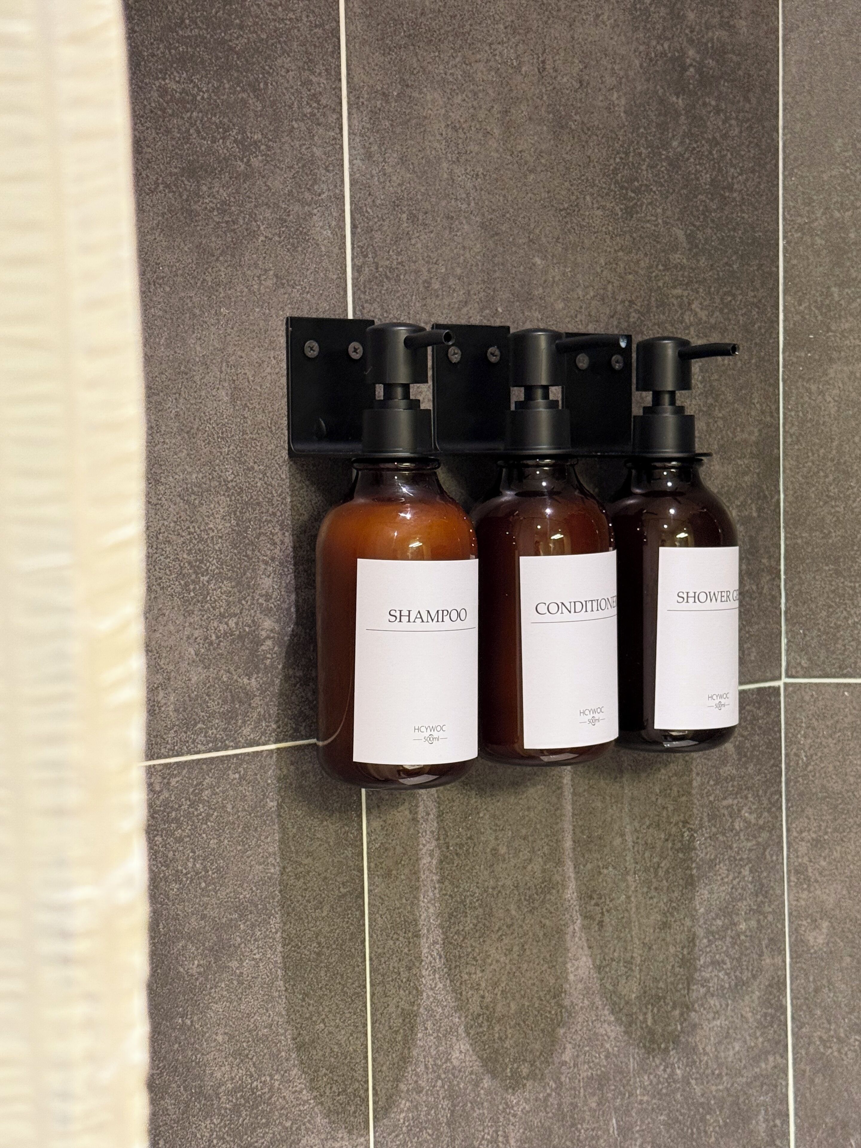 Three bottles of liquid products with pump dispensers, labeled 'SHAMPOO,' 'CONDITIONER,' and 'SHOWER GENTLE,' mounted on a tiled wall.
