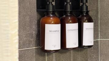 Three bottles of liquid products with pump dispensers, labeled 'SHAMPOO,' 'CONDITIONER,' and 'SHOWER GENTLE,' mounted on a tiled wall.