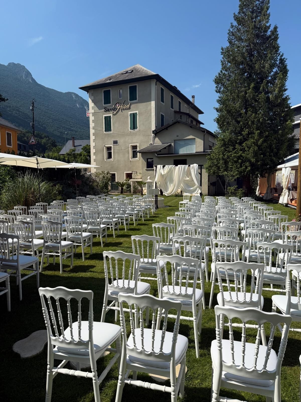 A hotel courtyard with rows of white chairs arranged for an event, a large building with a sign reading "Savo Hotel" in the background, and a mountain range visible beyond the property.
