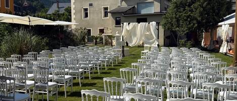 Une cour d’hôtel avec des rangées de chaises blanches disposées pour un événement, un grand bâtiment avec une enseigne indiquant « Savo Hotel » en arrière-plan, et une chaîne de montagnes visible au loin.