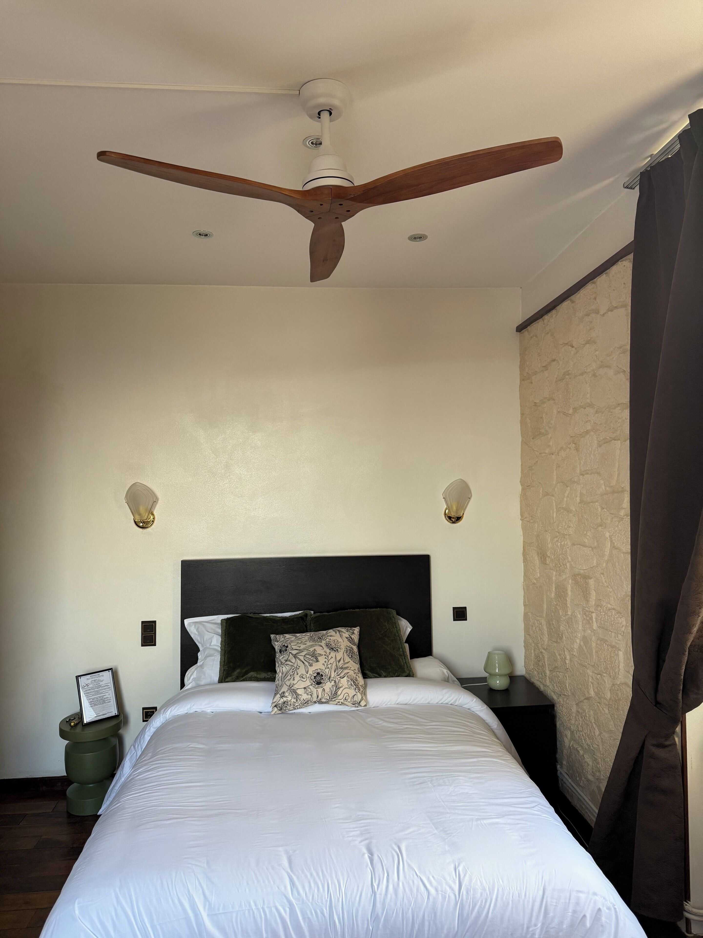 A bedroom with a large bed, a ceiling fan, two wall sconces, and a stone-textured wall.