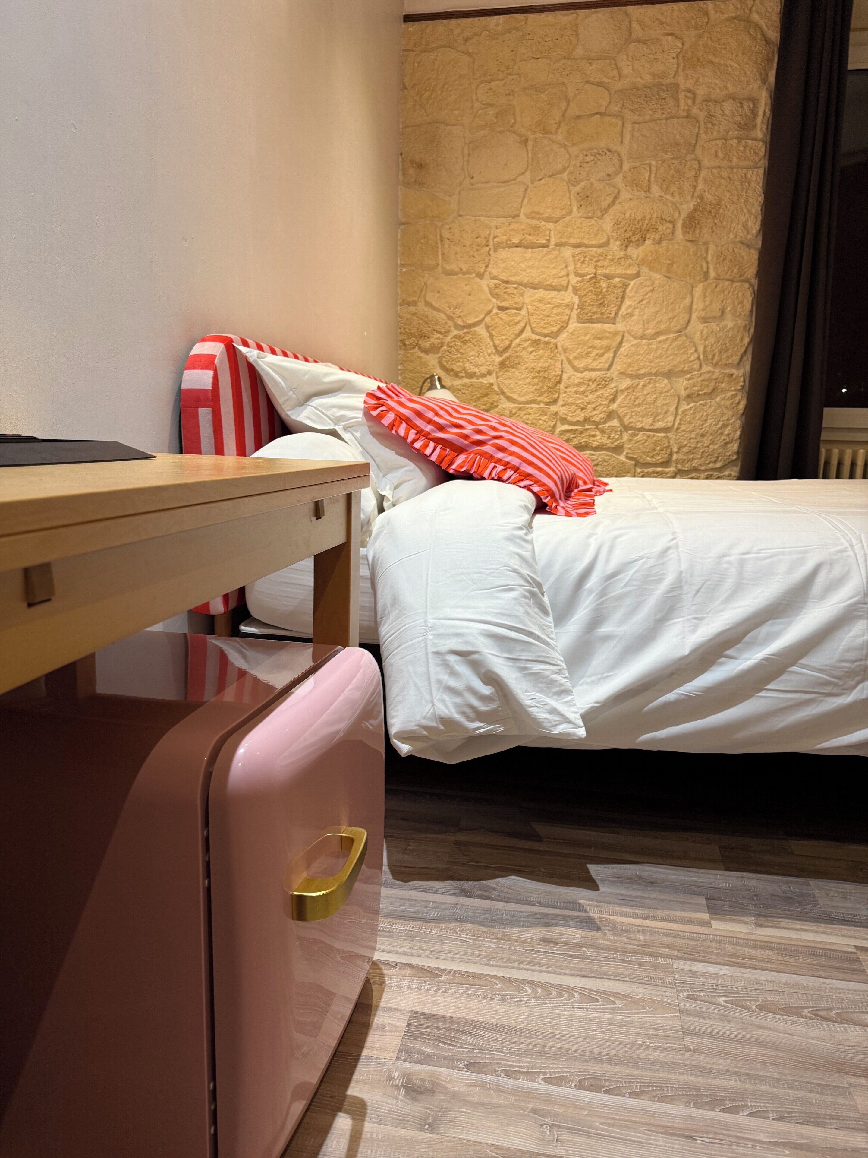 Una habitación de hotel con una cama, una maleta, una mesita de noche de madera y una pared de piedra.