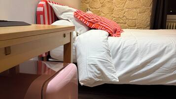 Una habitación de hotel con una cama, una maleta, una mesita de noche de madera y una pared de piedra.