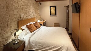 Een slaapkamer met een stenen muur, een houten plafondventilator, een bed met witte beddengoed, een nachtkastje met lamp en een aan de muur bevestigde televisie.