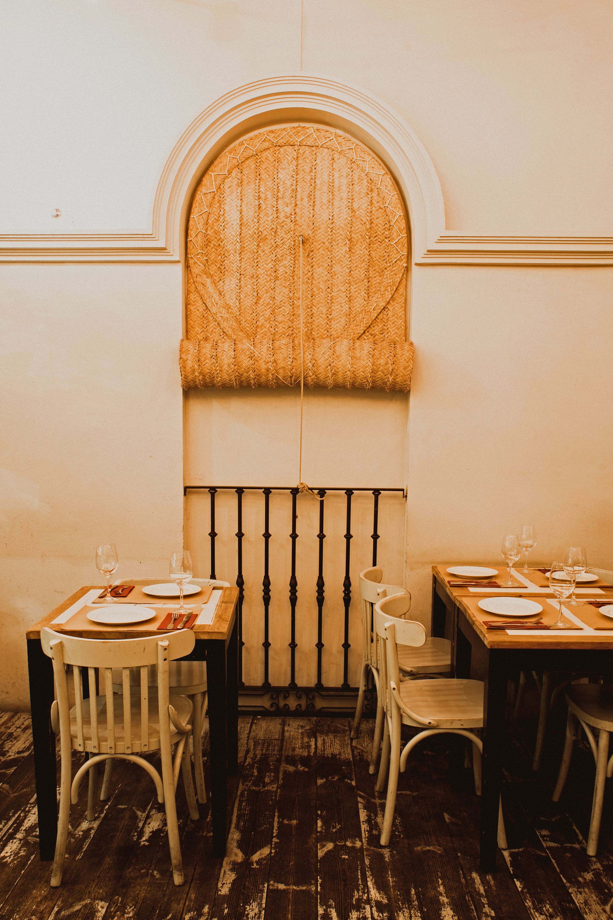 Un comedor con piso de madera rústica, sillas blancas y mesas dispuestas para dos, con copas de vino y platos.