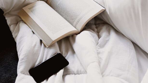 Una cama con un libro y un teléfono inteligente, una mesita con velas y una cámara, y una silla al fondo.