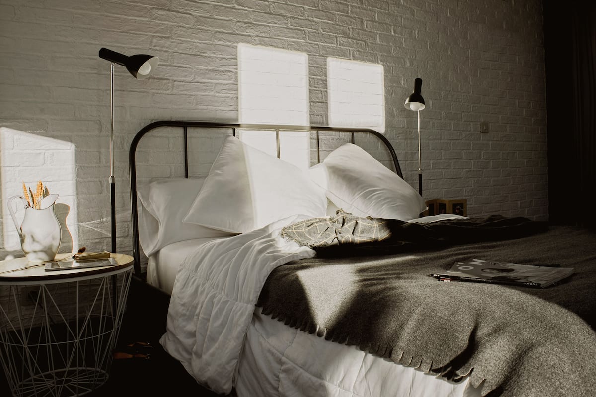 Ein ordentlich bezogenes Bett mit weißen Bettwäsche und einer grauen Tagesdecke, ein Nachttisch mit Lampe sowie eine Ziegelwand.
