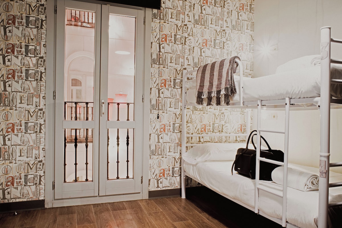 A bunk bed with a white mattress and a striped blanket, a black handbag on the lower bunk, and a patterned wallpaper with letters.