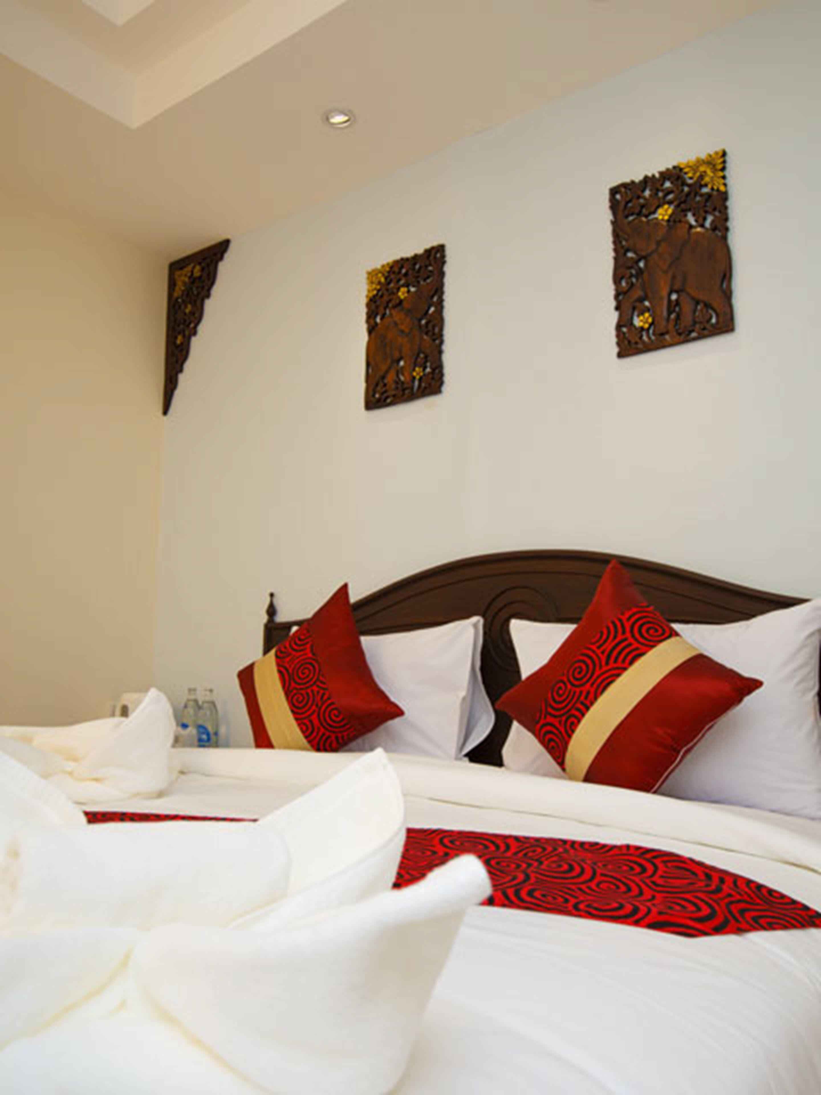 A neatly made bed with white linens and red and gold patterned pillows, a wooden headboard, and two wall-mounted artworks.