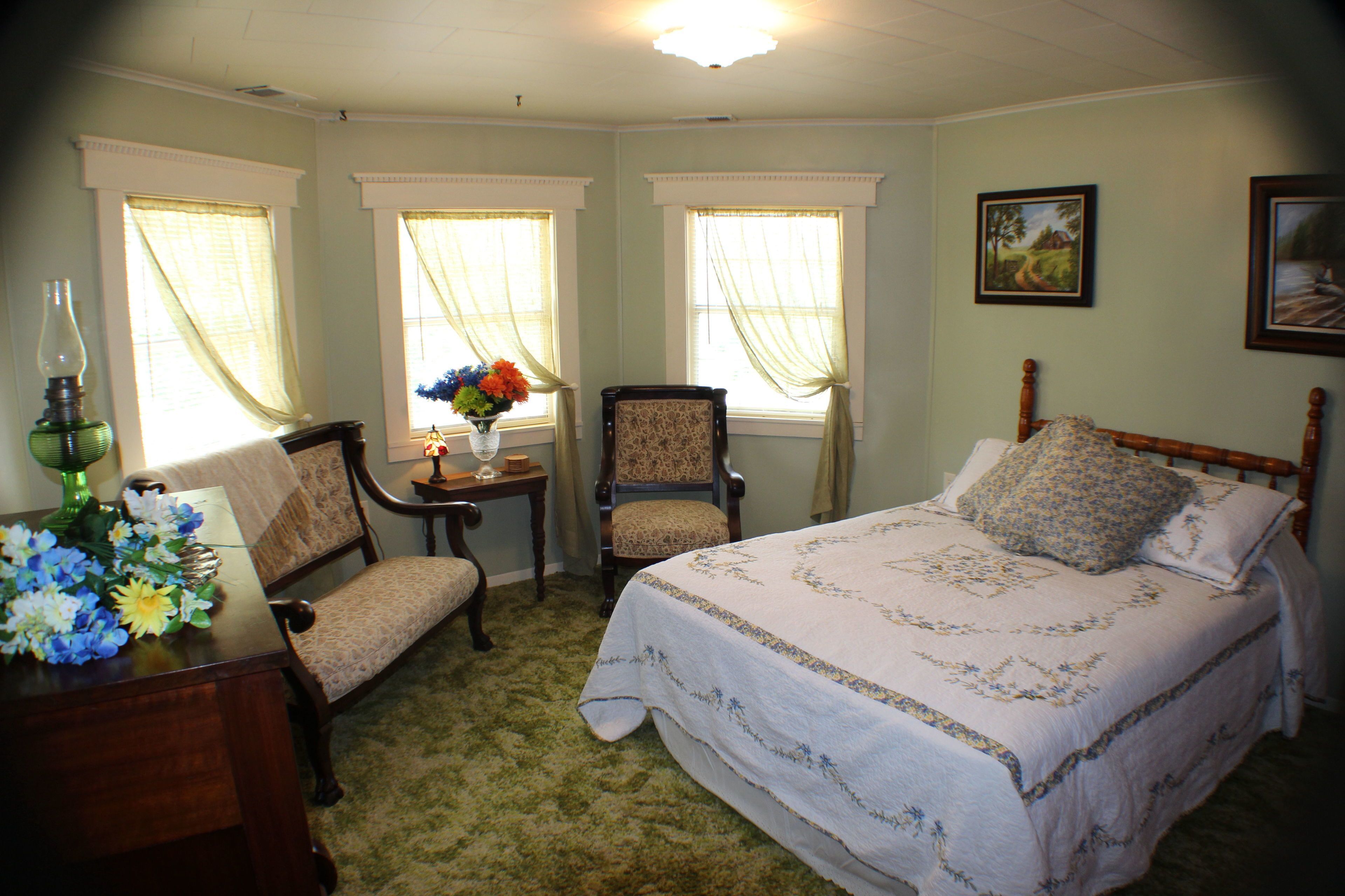 A bedroom with a large bed, two chairs, a small table with a vase of flowers, and two windows with curtains.