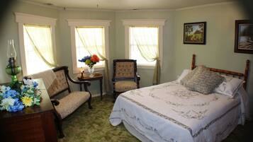 A bedroom with a large bed, two chairs, a small table with a vase of flowers, and two windows with curtains.