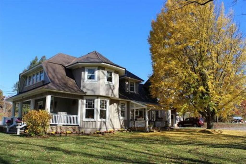 A two-story house with a wrap-around porch, a gabled roof, and a large tree with yellow leaves in the yard.