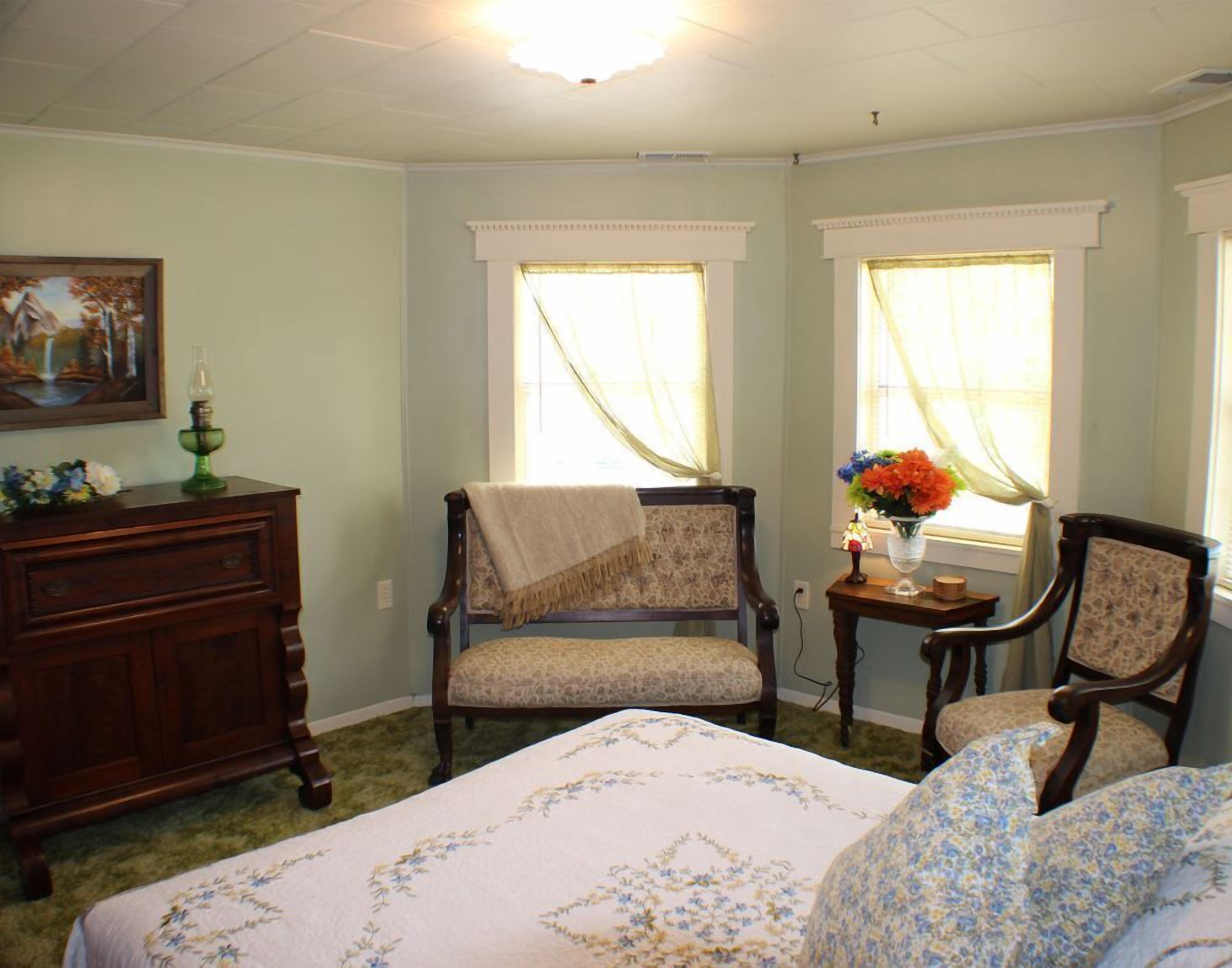 A room with a bed, two chairs, a dresser, and two windows with curtains.