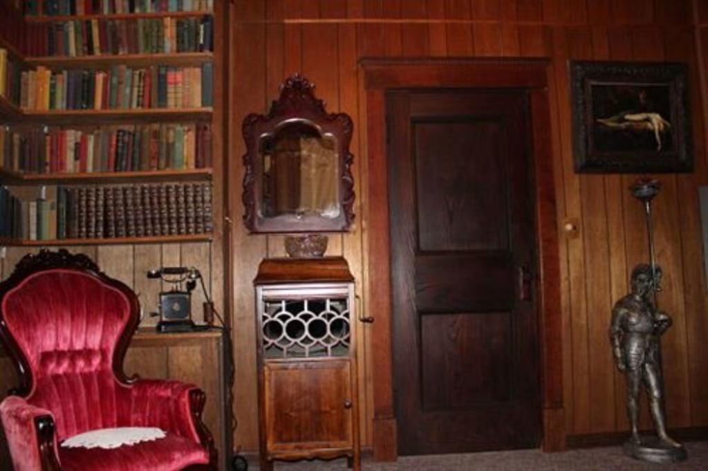 A room with wooden paneling, a bookshelf, a red armchair, a small cabinet, a door, and a statue.