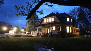Une maison à deux étages avec un porche, une balançoire et un jardin bien éclairé à l’avant, au crépuscule.