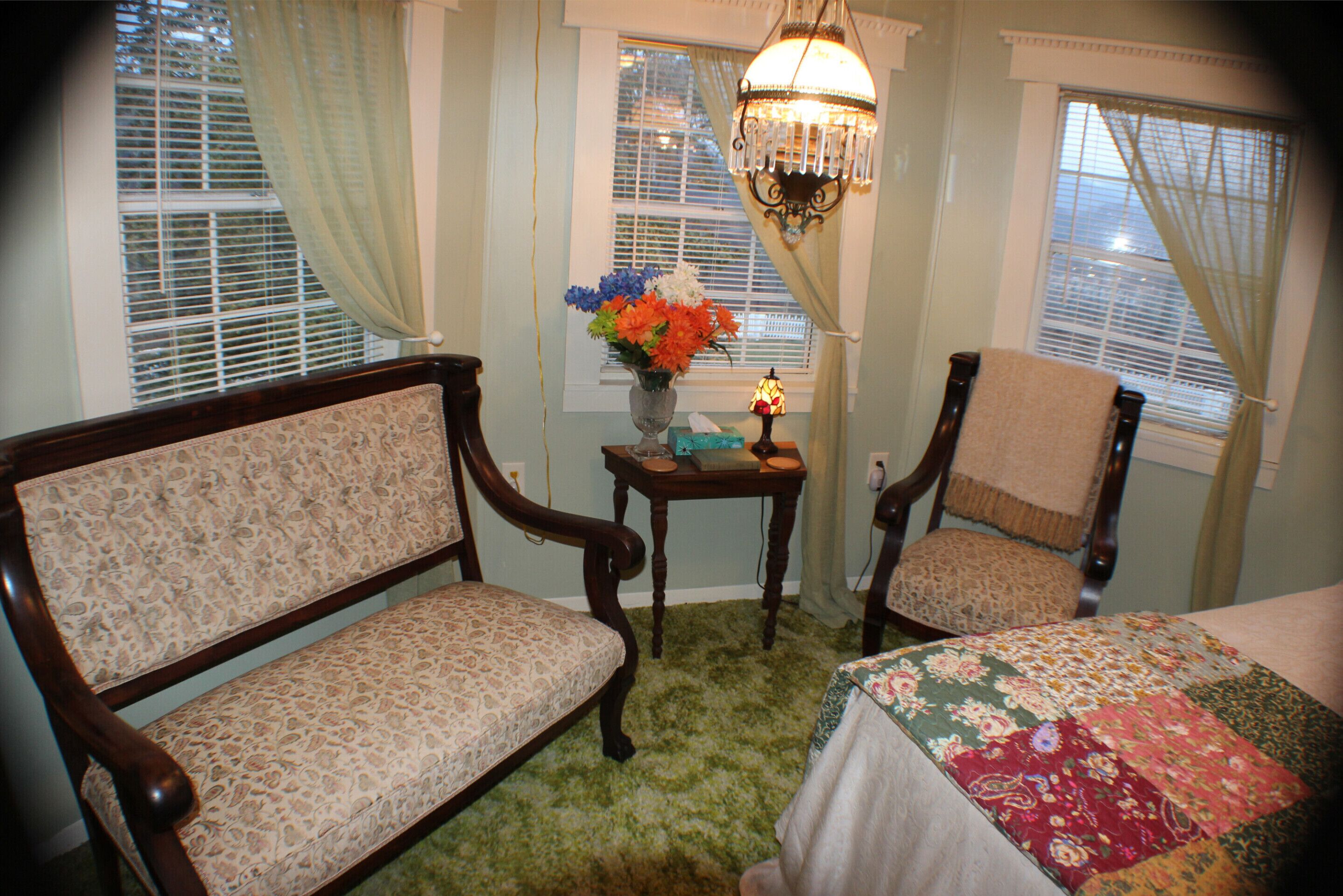 A room with two wooden chairs, a small table with a vase of flowers, and a bed with a floral bedspread.