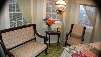 A room with two wooden chairs, a small table with a vase of flowers, and a bed with a floral bedspread.