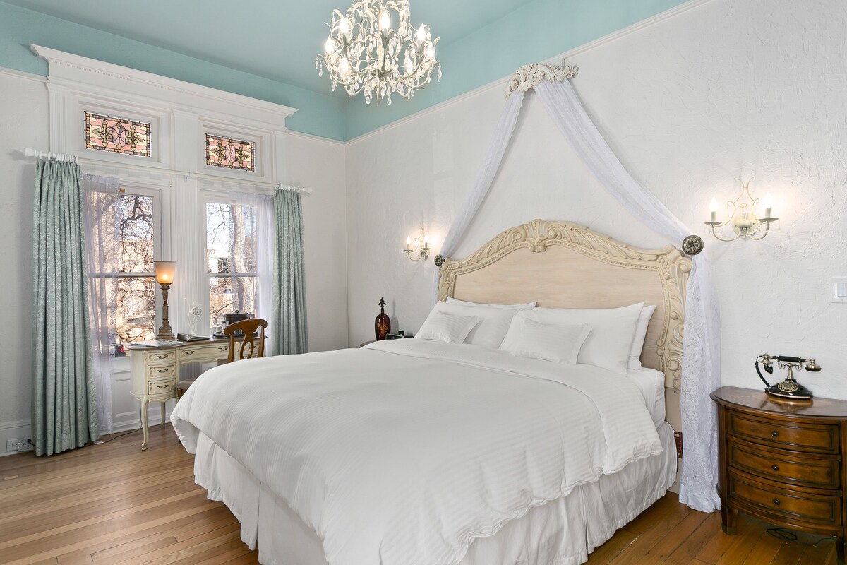 A bedroom with a four-poster bed, a wooden nightstand, a desk with a lamp, and a chandelier.