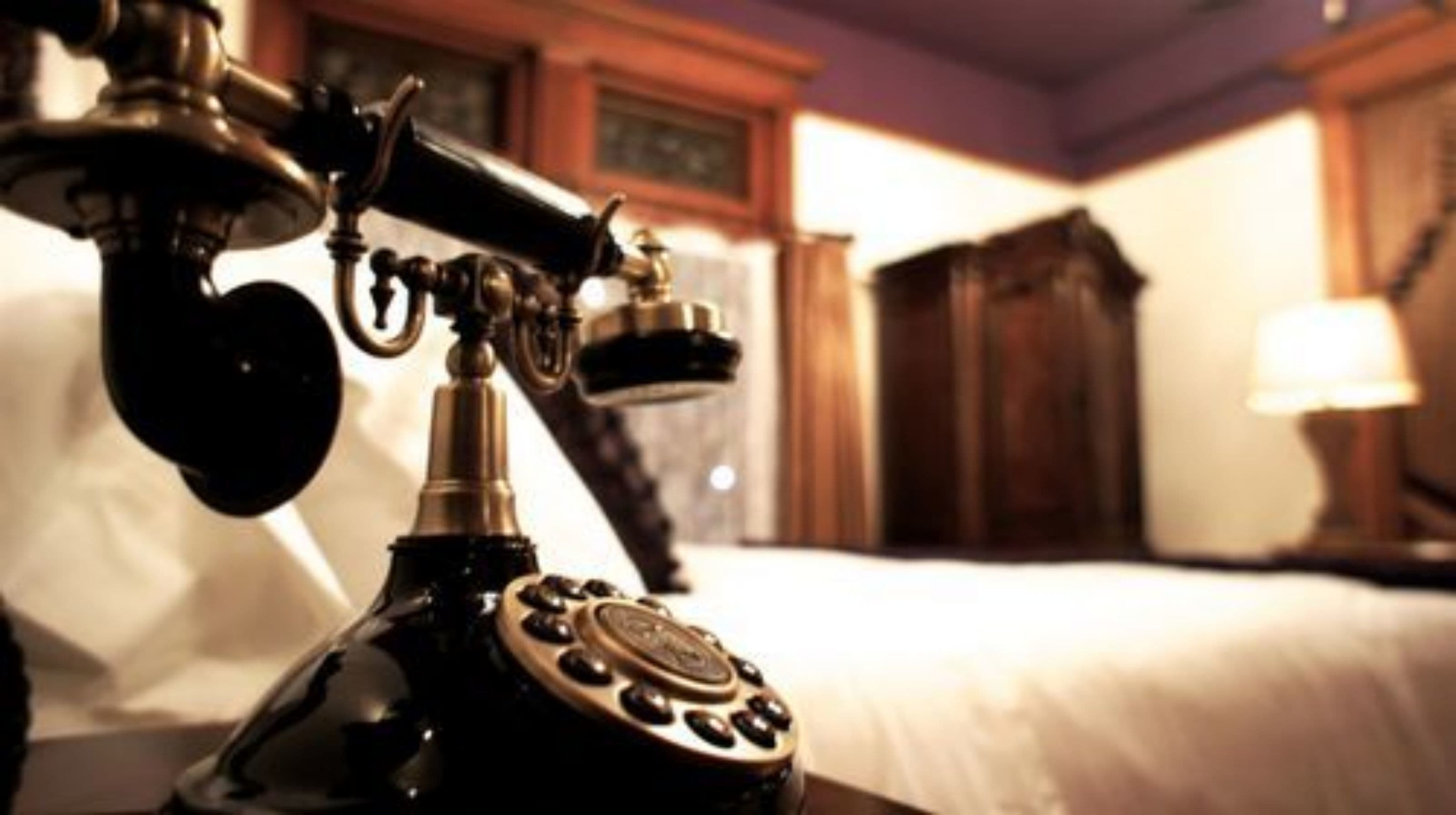 A vintage telephone on a bedside table in a hotel room with a bed and a wooden cabinet in the background.