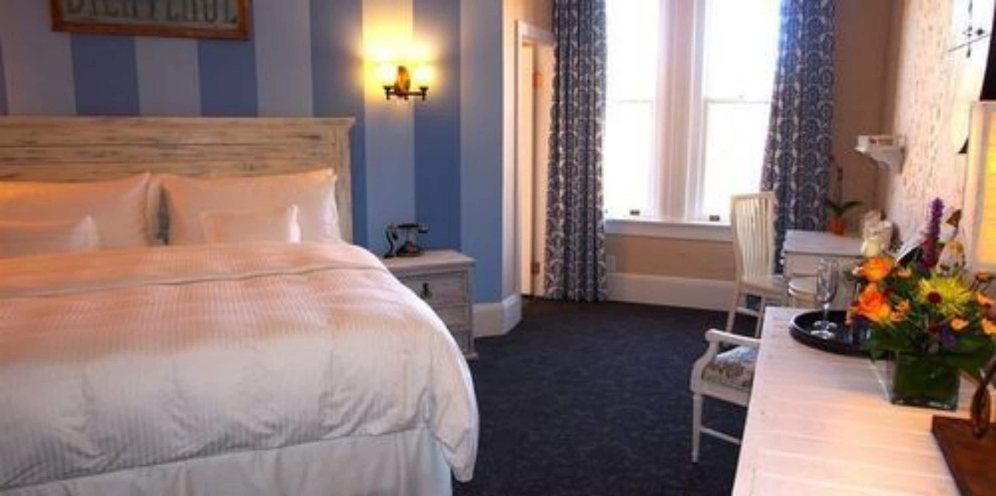 A hotel room with a large bed, a bedside table, a chair, a desk with a vase of flowers, and a window with curtains.