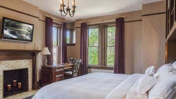 A hotel room with a fireplace, a desk, a bed, and large windows with curtains.