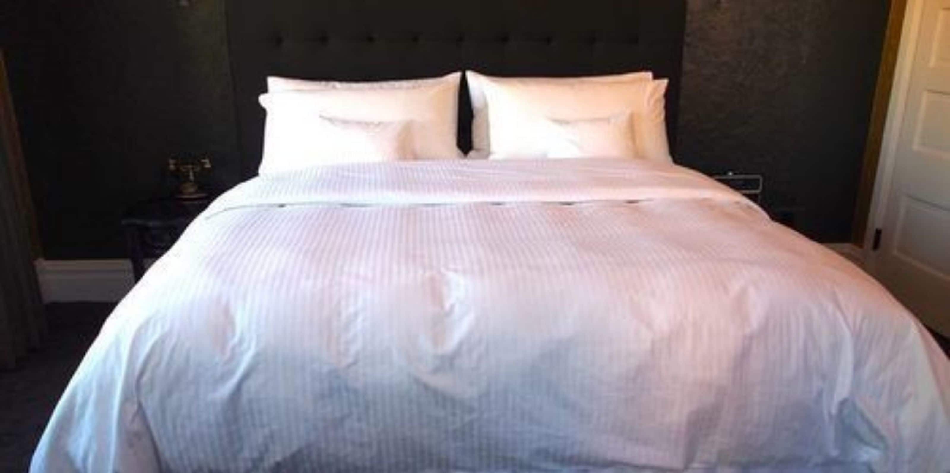Un lit bien fait, avec du linge de lit blanc, dans une pièce aux murs sombres et dotée d’une tête de lit capitonnée.