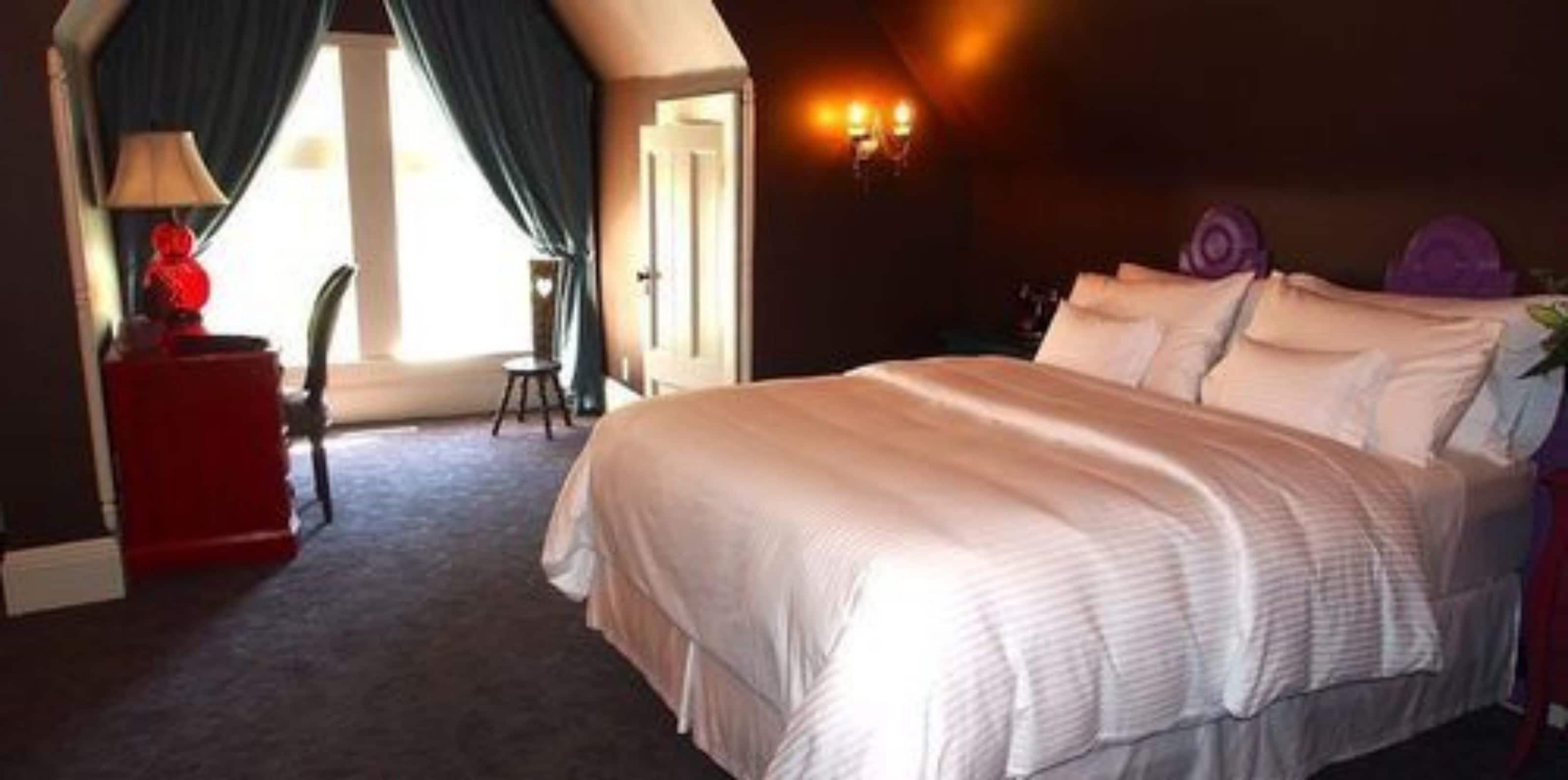 A hotel room with a large bed, a red side table, a lamp, and a view of the outdoors.