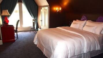 A hotel room with a large bed, a red side table, a lamp, and a view of the outdoors.