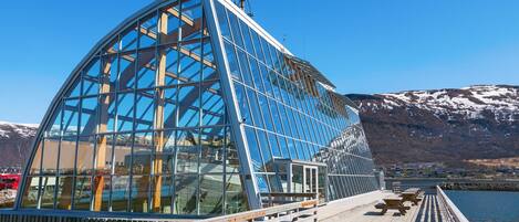 A modern glass structure with a wooden deck and benches, overlooking a body of water and snow-capped mountains.
