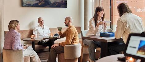 A group of people seated in a modern lounge area with round tables, a large window with a view, and a wall-mounted artwork.