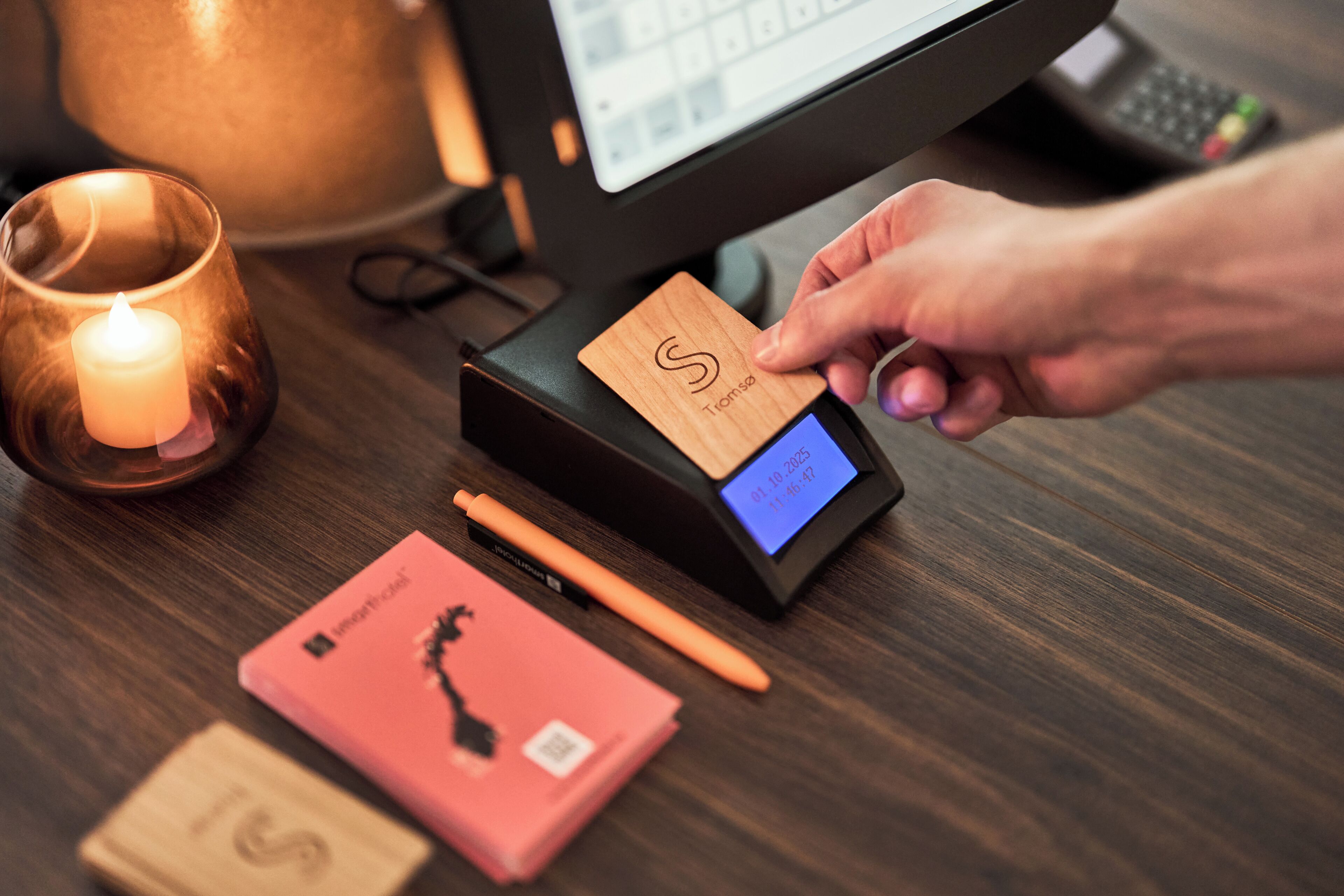 Una tarjeta de madera siendo escaneada en un terminal de punto de venta, con una pantalla digital, una vela y una libreta rosa sobre una superficie de madera.