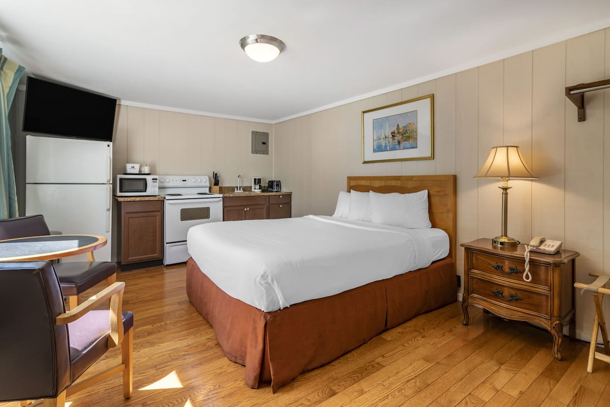 A hotel room with a bed, bedside table, lamp, and a kitchenette area.