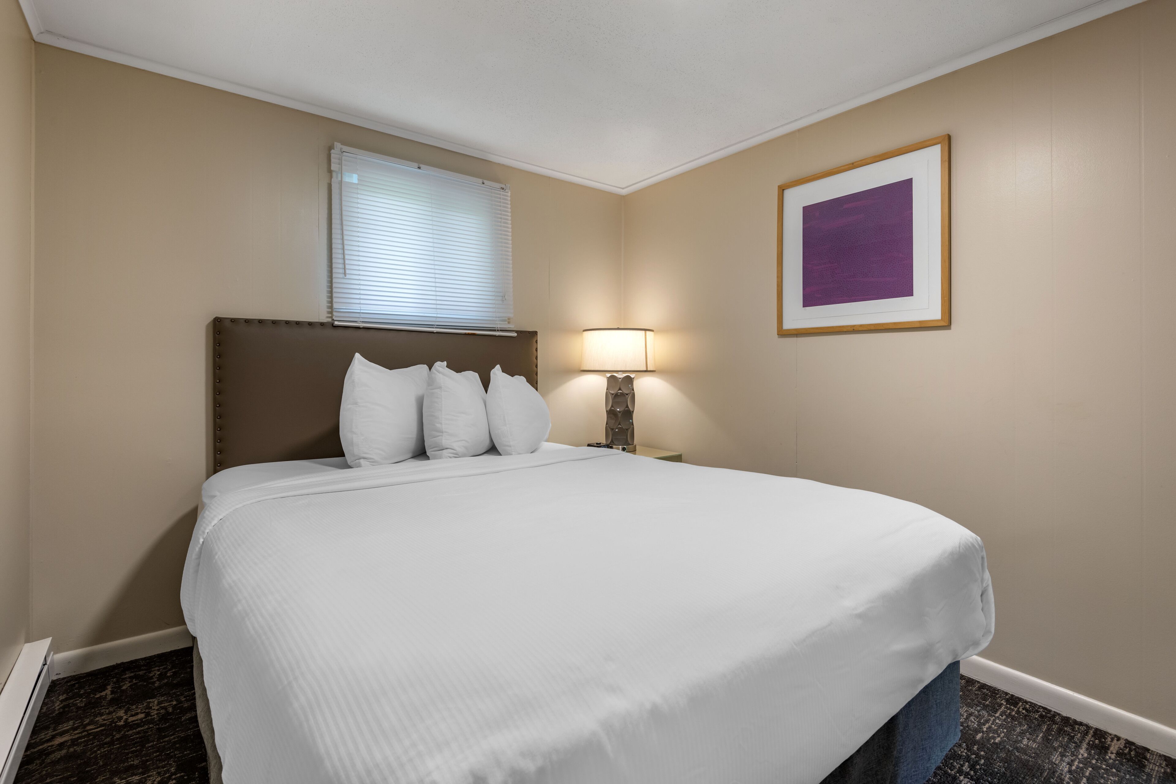Une chambre d’hôtel avec un grand lit, une lampe de chevet, une fenêtre avec des stores et un tableau encadré au mur.
