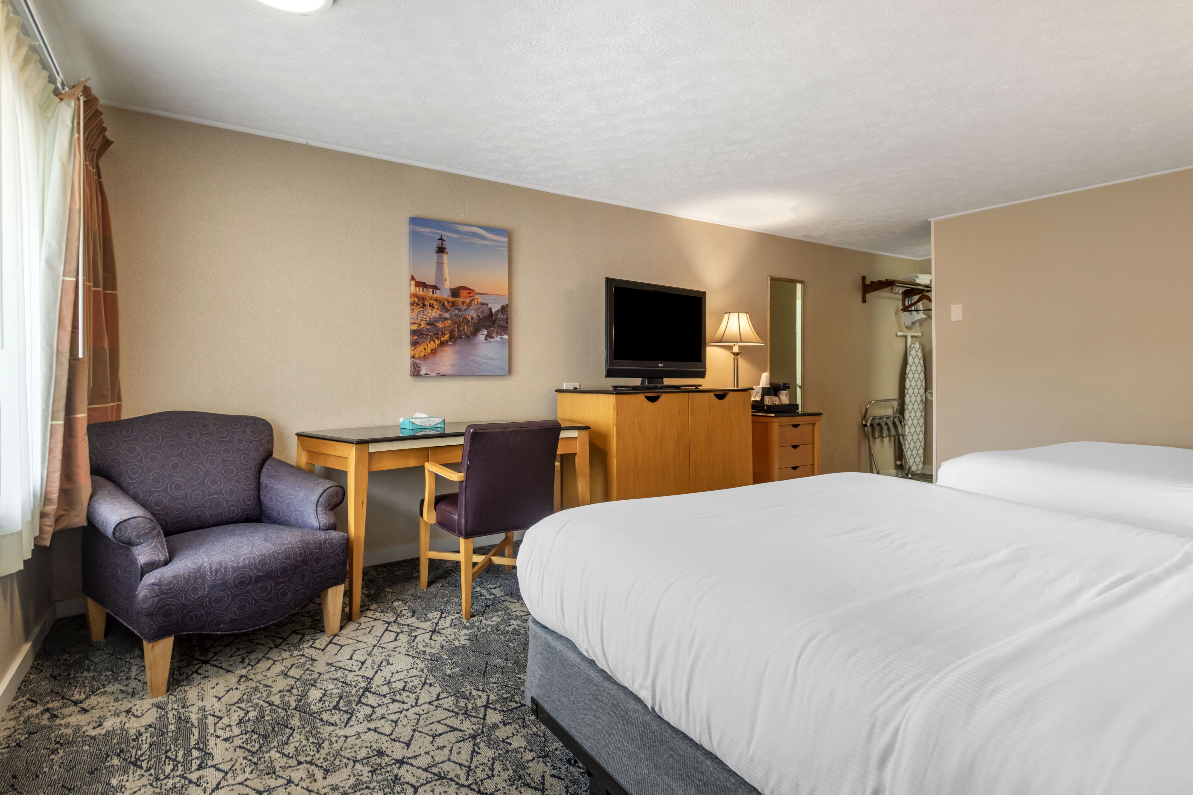 A hotel room with a bed, a desk, a chair, a television, and a painting on the wall.