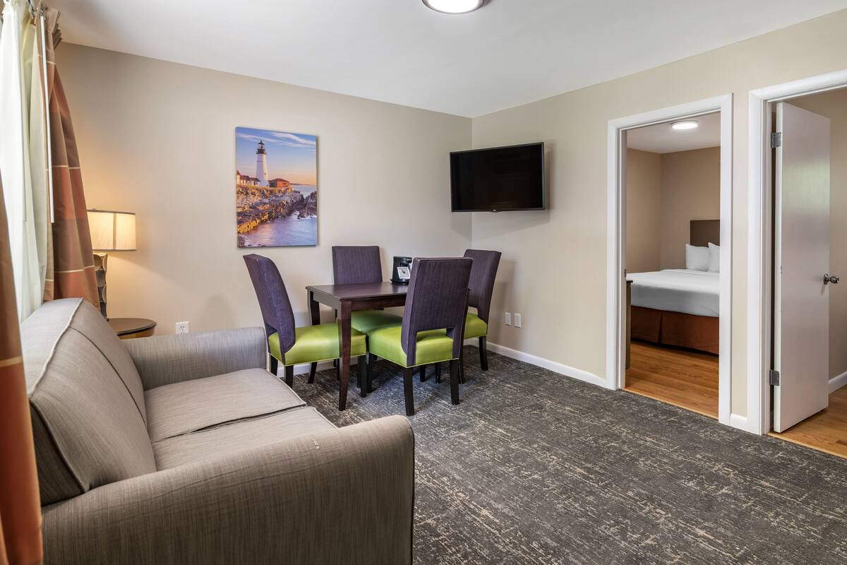 A hotel room with a sofa, dining table, chairs, a flat-screen TV, and a bed.
