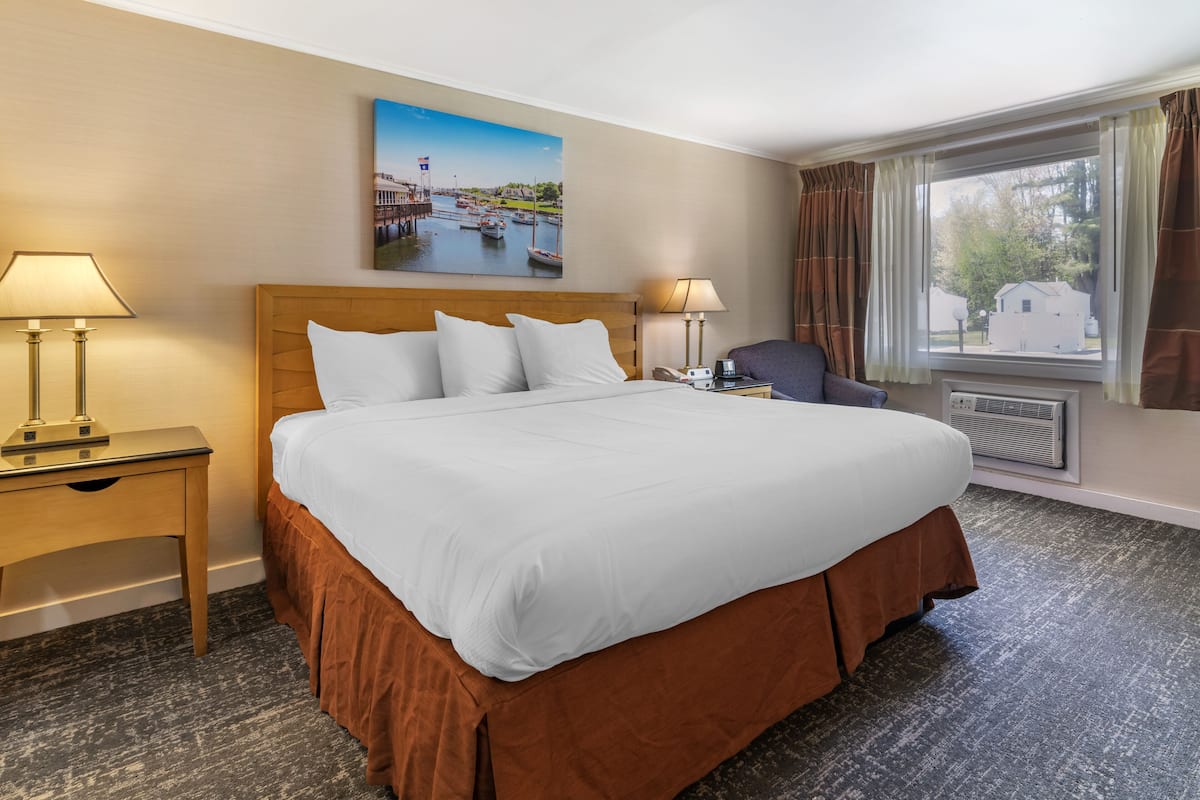 A hotel room with a large bed, two bedside lamps, a nightstand, a chair, a painting on the wall, and a window with curtains.