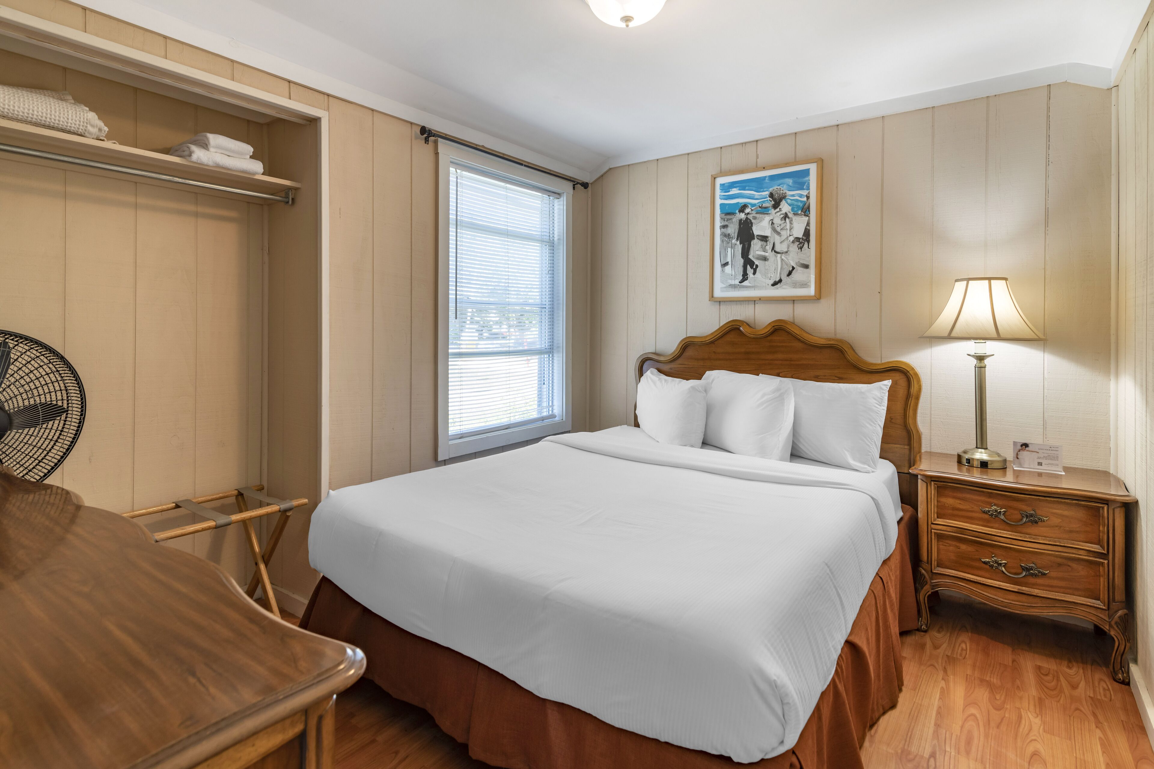 Un dormitorio ordenado con una cama de madera, una mesita de noche con lámpara, una ventana con persianas y un ventilador.