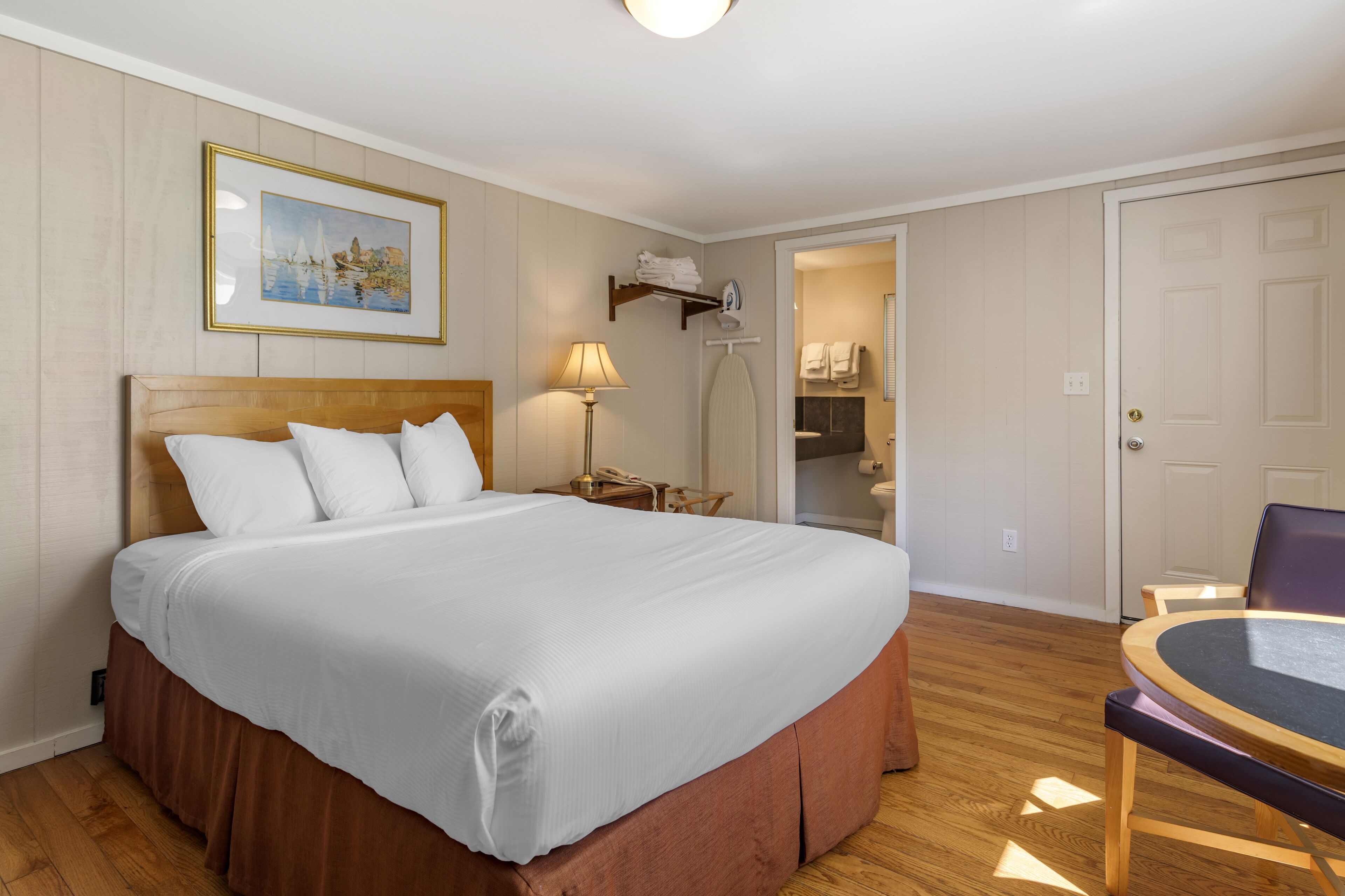 Una habitación de hotel ordenada con una cama grande, una mesita de noche con lámpara, una silla y una pequeña mesa con superficie de vidrio.