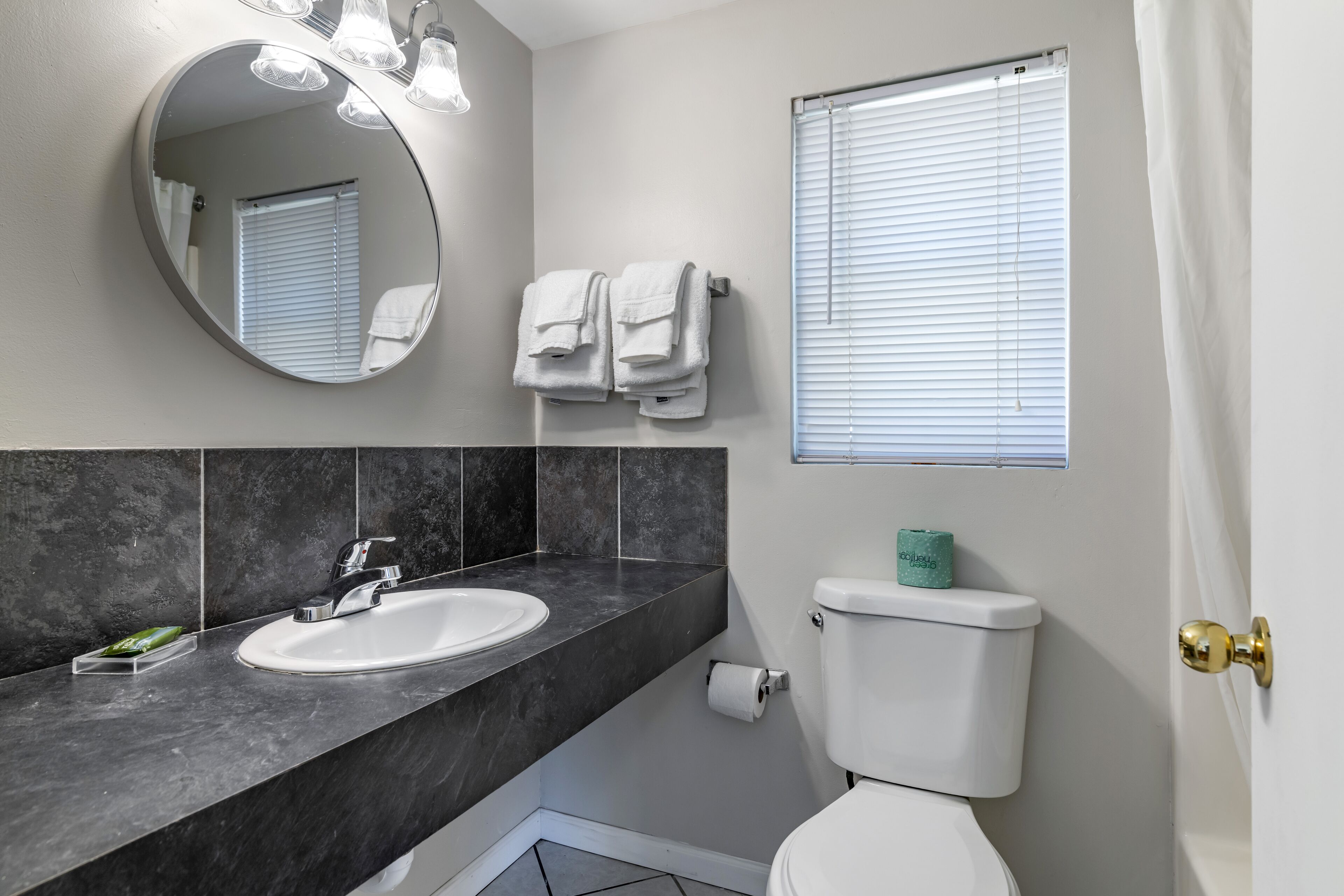 A bathroom with a round mirror, a sink, a toilet, and a window with blinds.