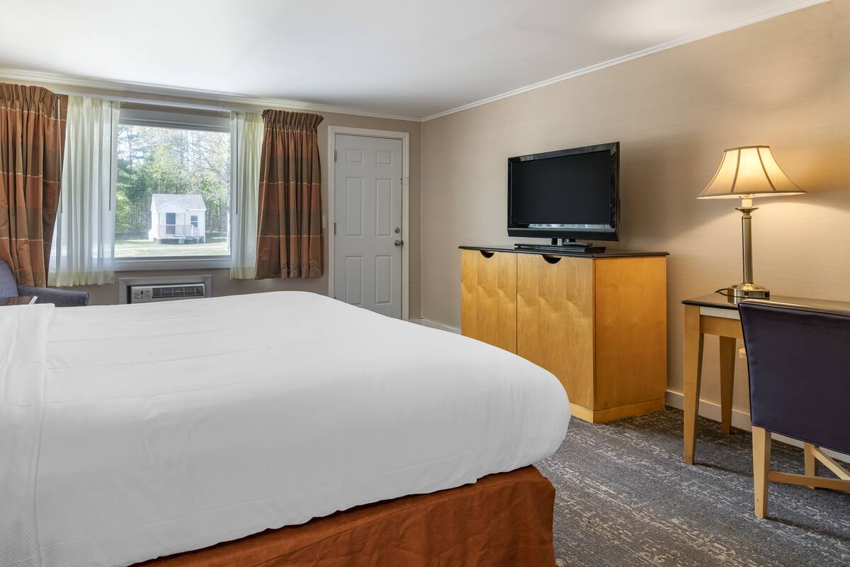 A hotel room with a large bed, a television on a wooden cabinet, a desk with a lamp, and a window with curtains.