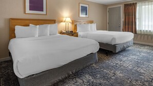 A hotel room with two beds, a nightstand with a lamp, framed artwork on the walls, and a patterned carpet. - Anchor Inn and Cottages Wells-Ogunquit (Wells)