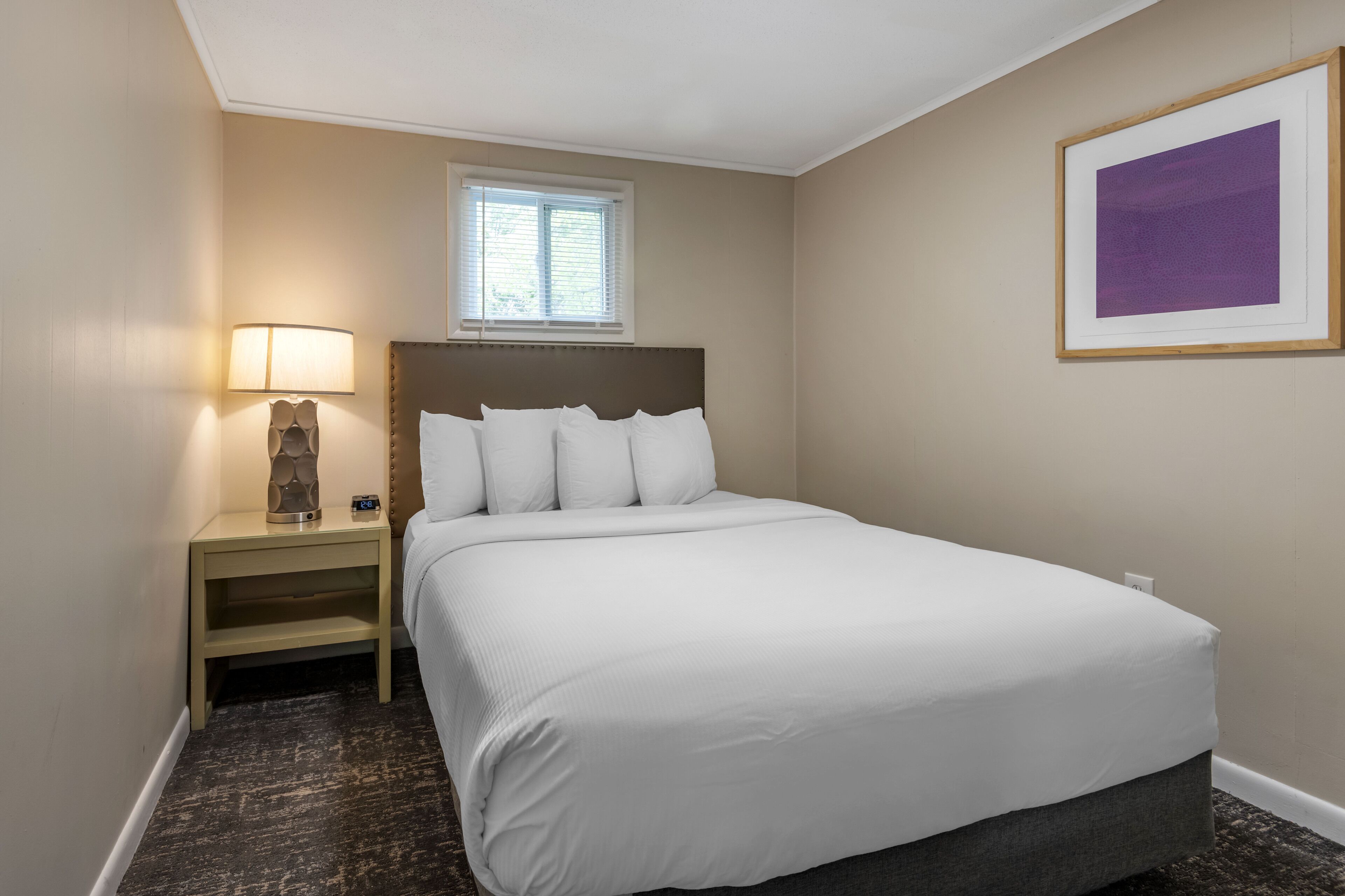 Habitación de hotel ordenada con cama, mesita de noche, lámpara, ventana y un cuadro en la pared.