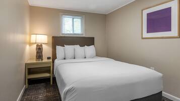 Habitación de hotel ordenada con cama, mesita de noche, lámpara, ventana y un cuadro en la pared.