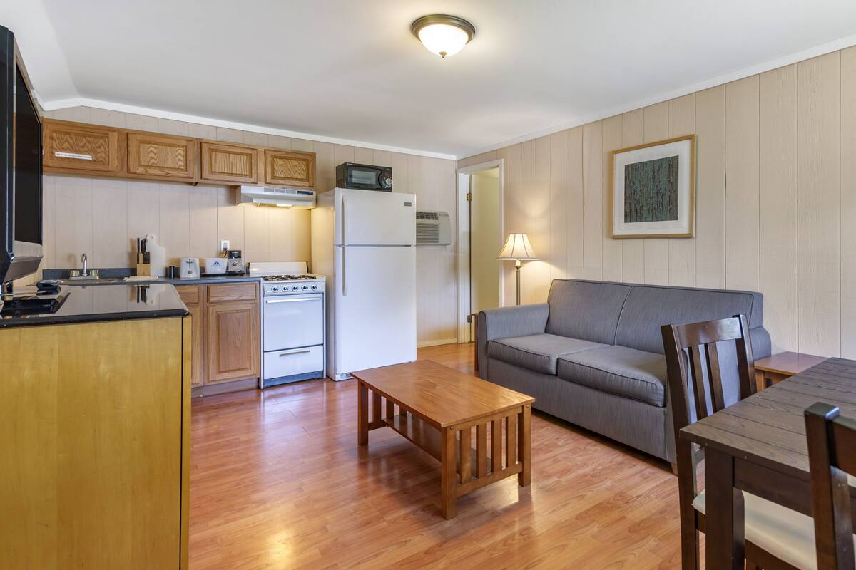 A compact kitchen with wooden cabinets, a refrigerator, a microwave, and a stove. There is a gray sofa, a wooden coffee table, and a dining table with chairs.