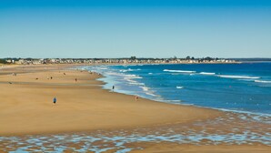 A wide beach with people walking along the shore, a clear blue sky, and buildings in the distance. - Anchor Inn and Cottages Wells-Ogunquit (Wells)