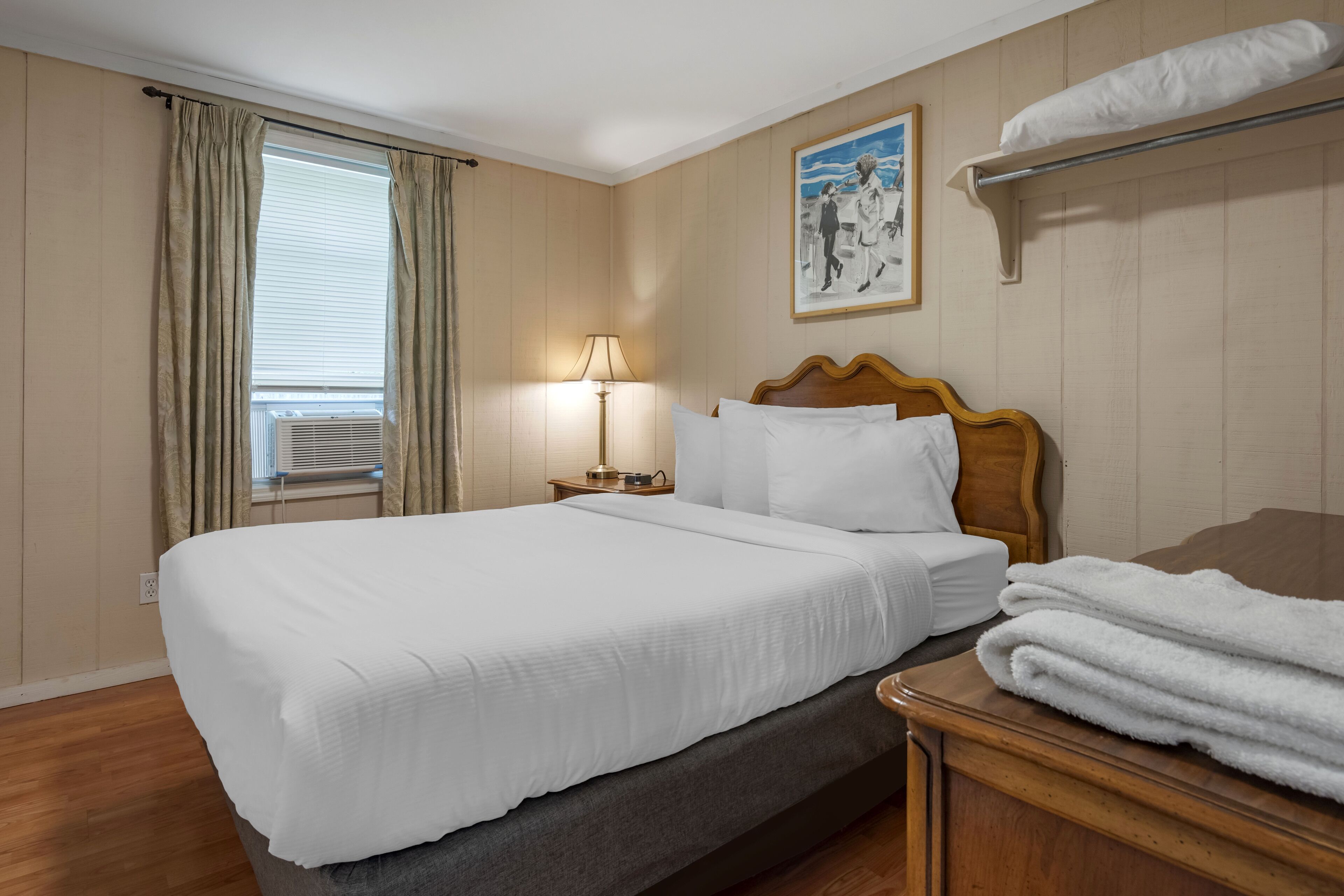 Una cama bien hecha con sábanas blancas, una lámpara de noche, un cabecero de madera y una ventana con cortinas.