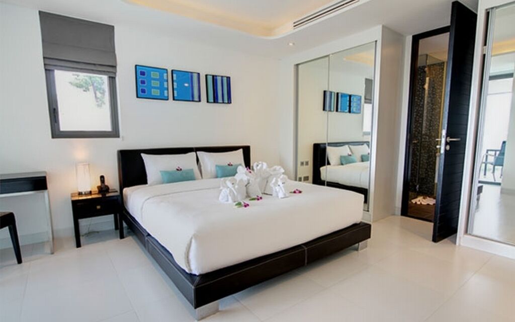 A modern hotel bedroom with a large bed, a bedside table, a mirror, and a sliding door to a balcony.