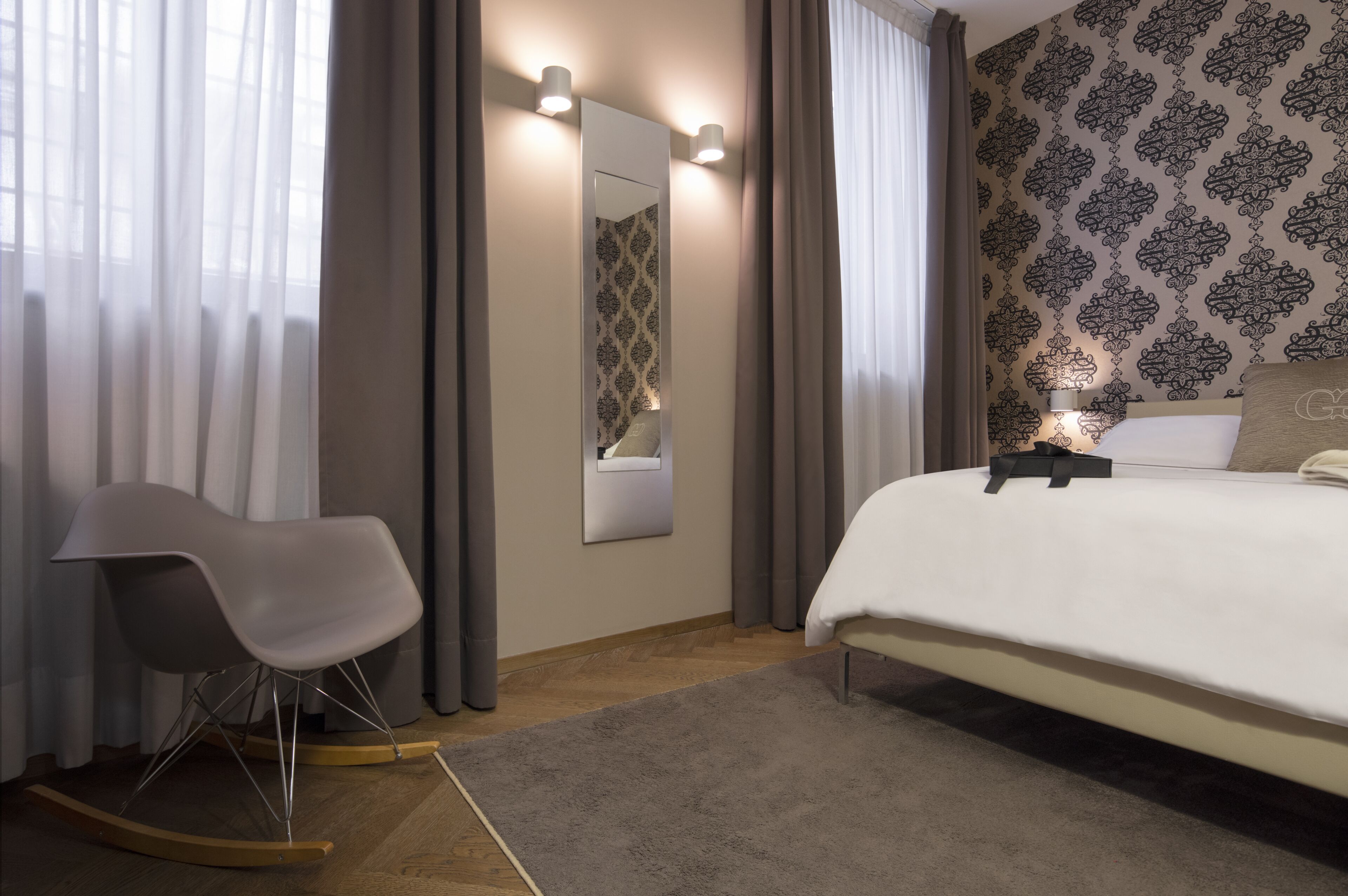 A modern bedroom with a large bed, a rocking chair, a mirror, and patterned wallpaper.