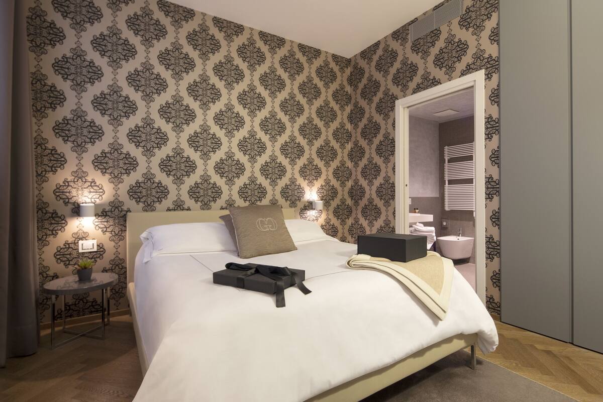 Una camera da letto ordinata con un letto grande, comodini con lampade, un tavolino rotondo con una pianta e un bagno visibile attraverso una porta aperta.